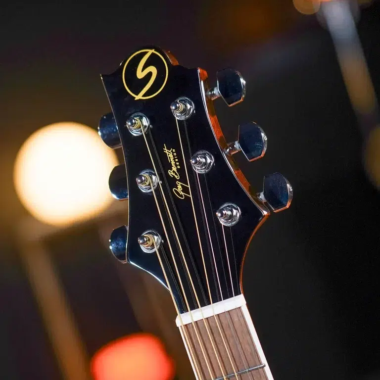 Close-up of a black guitar headstock with six chrome tuning pegs and six steel strings, gold logo on the headstock, warm stage lighting in the background. ขายราคาพิเศษ