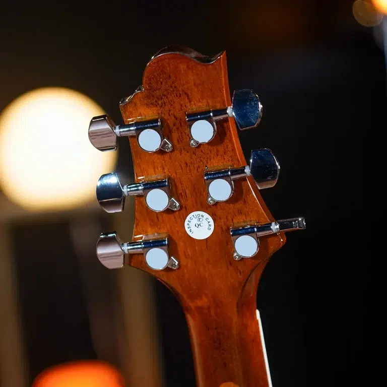 Close-up of a wooden guitar headstock with chrome tuning pegs and a white QC sticker. ขายราคาพิเศษ