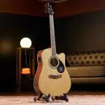 Acoustic guitar on a stand in a dim room with a round lamp and tufted sofa in the background. ขายราคาพิเศษ
