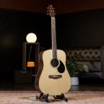 Acoustic guitar on a stand in a warmly lit living room, with a lamp, sofa, and a potted plant in the background. ลดราคาพิเศษ