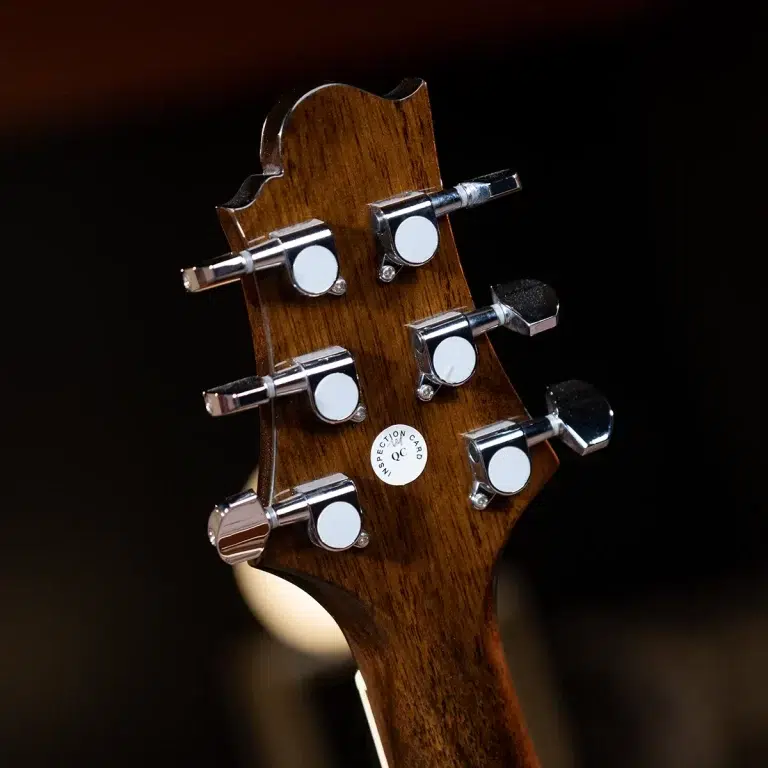 Close-up of a wooden guitar headstock with chrome tuning pegs and a QC inspection sticker on a dark background. ขายราคาพิเศษ