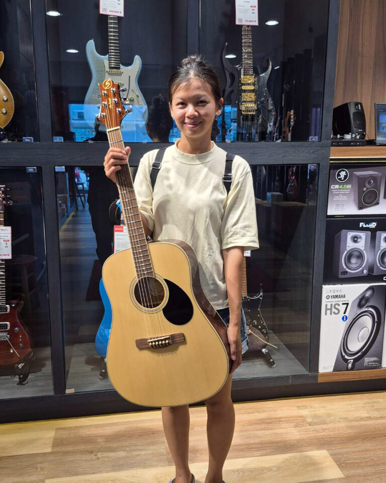 Young woman in a beige shirt holding an acoustic guitar in a music shop display of guitars behind her.