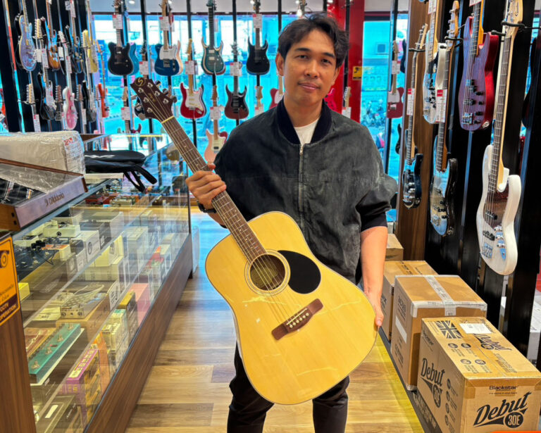 Man in a music shop holding an natural wood acoustic guitar with rows of electric guitars on the wall behind him.