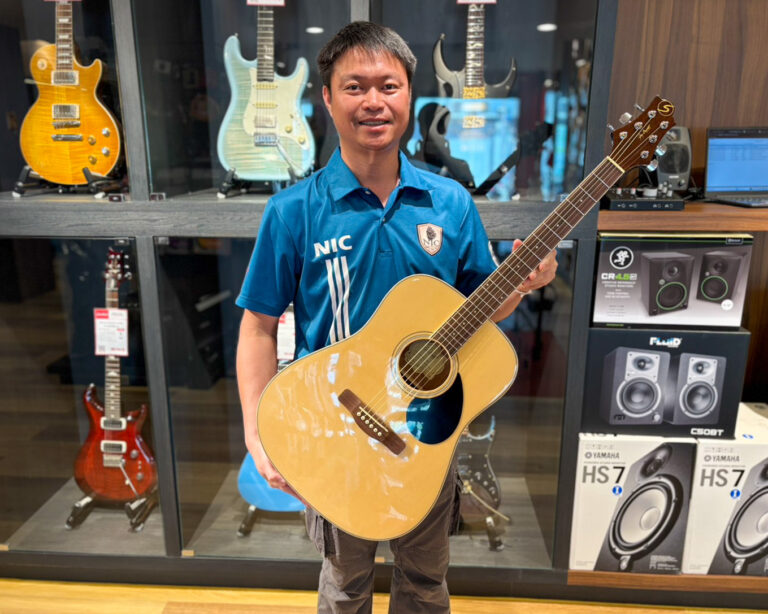 Man in a blue polo holds an acoustic guitar in a music store showroom.