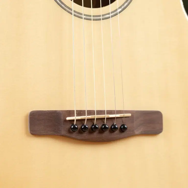 Close-up of an acoustic guitar bridge with six strings anchored by black bridge pins on a light wood body. ขายราคาพิเศษ