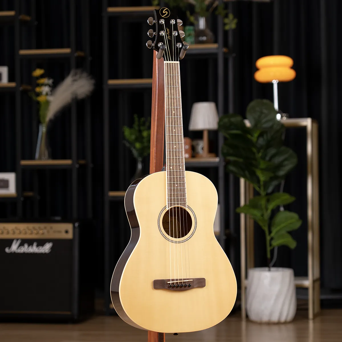 Acoustic guitar on a stand in a cozy studio with a Marshall amp and plants in the background.