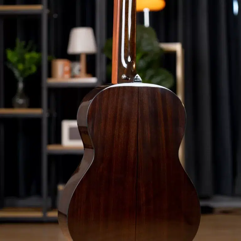 Back of a polished dark‑wood acoustic guitar in a cozy room with blurred shelves and a lamp in the background, seen from behind. ขายราคาพิเศษ