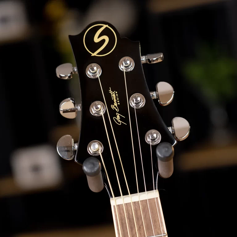 Close-up of a black guitar headstock with six chrome tuning pegs, a gold circular emblem at the top, and a cursive brand name along the headstock with strings running to the nut. ขายราคาพิเศษ