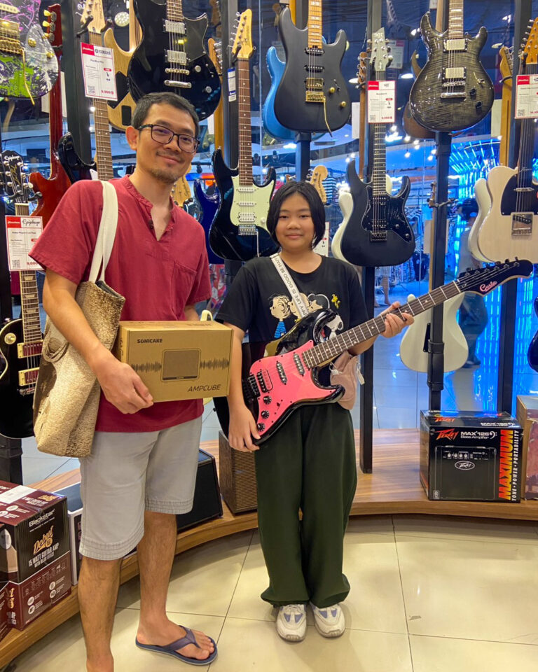 Two people stand in a guitar shop: a man in red shirt holding a boxed amp and a bag, and a girl with a pink electric guitar on loan or purchase.