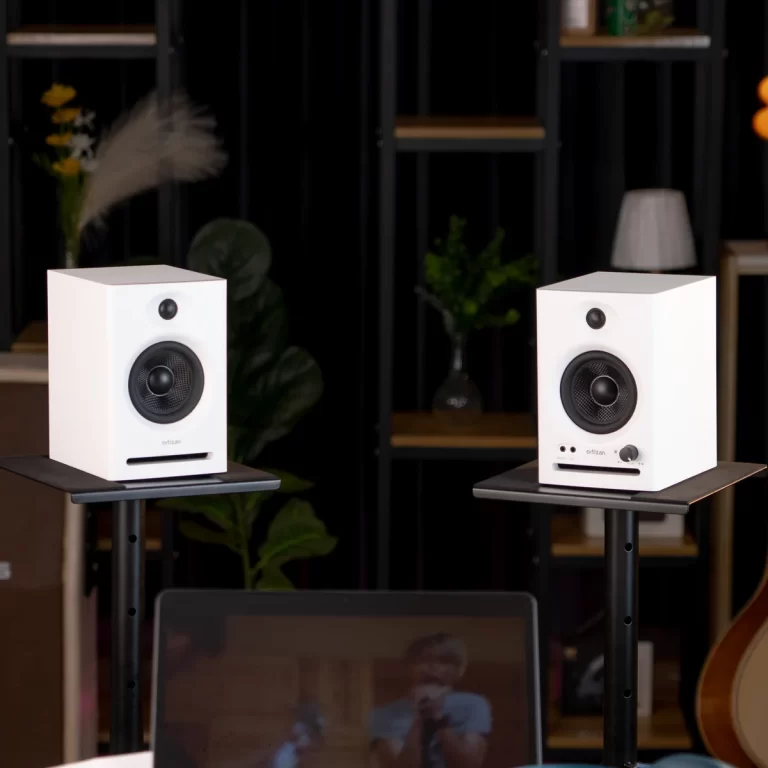 Two white Ortizan speakers on black stands in a home studio, with shelves and plants in the background. ขายราคาพิเศษ