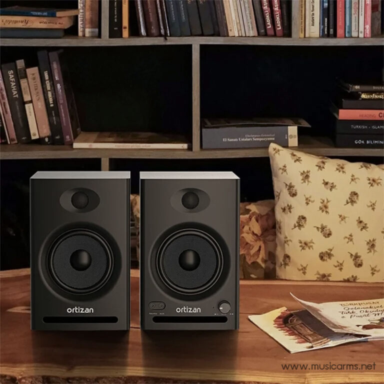 Two gray Ortizan bookshelf speakers sit on a wooden table in front of a dark bookshelf filled with books; a floral cushion and scattered papers are nearby, creating a cozy living-room vibe. ขายราคาพิเศษ