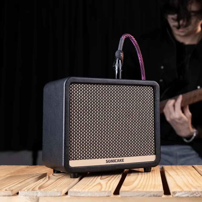 Small guitar amplifier on a wooden table with a musician playing in the background, pink cable plugged in ขายราคาพิเศษ