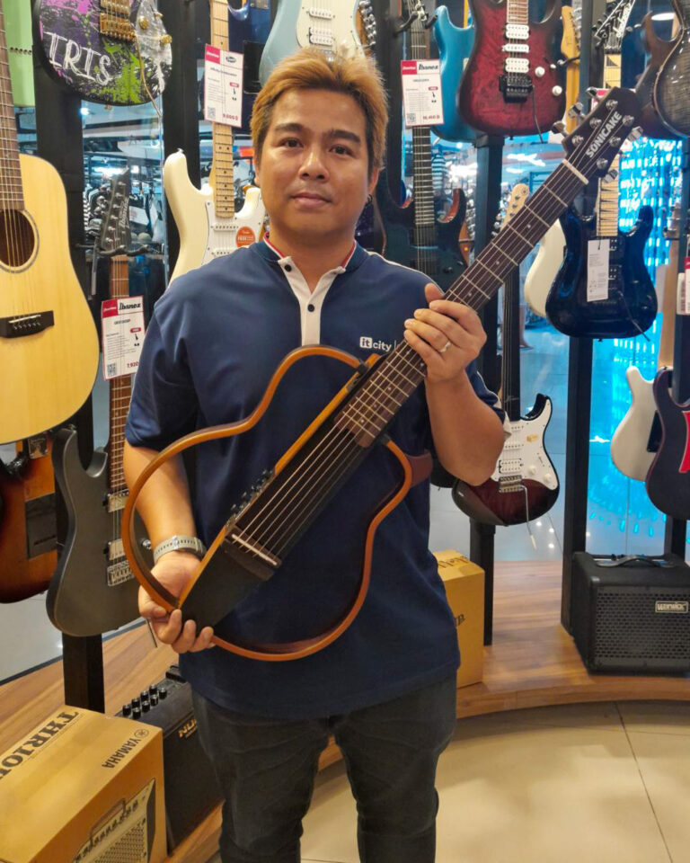 Man in a music store holding a curved-body guitar with a long neck, surrounded by hanging guitars and amps in the background.