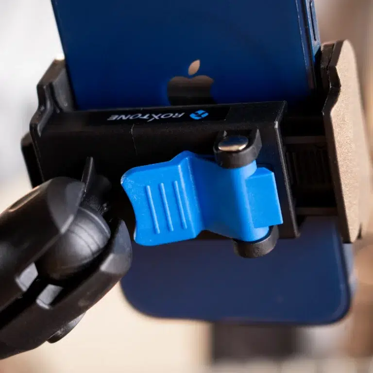 Close-up of a blue mobile phone clamp gripping a blue iPhone in a mount, showing the securing lever and arm. ขายราคาพิเศษ