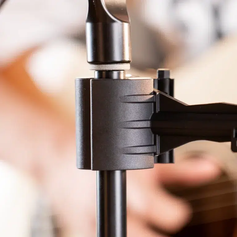 Close-up of a black microphone stand clamp securing a vertical pole, with a blurred guitarist in the background. ขายราคาพิเศษ
