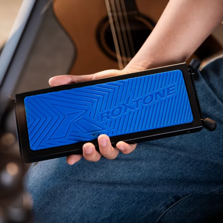 Person holding a black rectangular device with a bright blue textured panel in front, guitar in the background ขายราคาพิเศษ