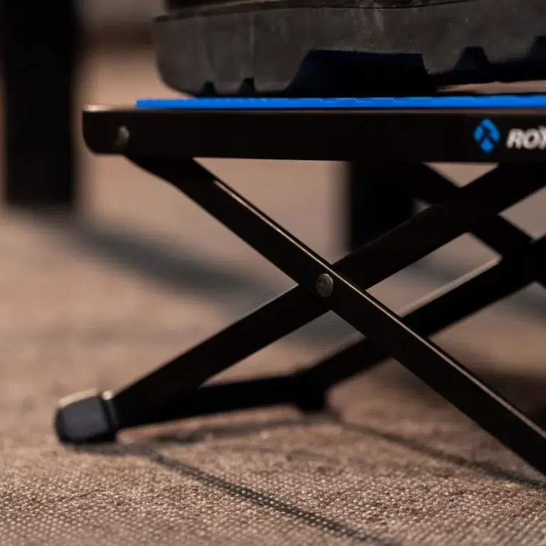 Close-up of a black folding chair frame with X-shaped legs on a carpeted floor, blue seat edge visible above the frame. ขายราคาพิเศษ
