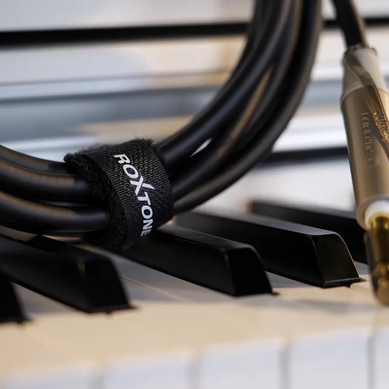 Close-up of a black audio cable with a Velcro strap lying across piano keys, with a 1/4-inch plug nearby. It emphasizes connection/setup for a keyboard or instrument. ขายราคาพิเศษ