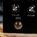 Close-up of a black guitar effects pedal front panel with two round knobs labeled 'DRIVE LEVEL' and 'DRIVE GAIN' and a metallic footswitch below. ขายราคาพิเศษ