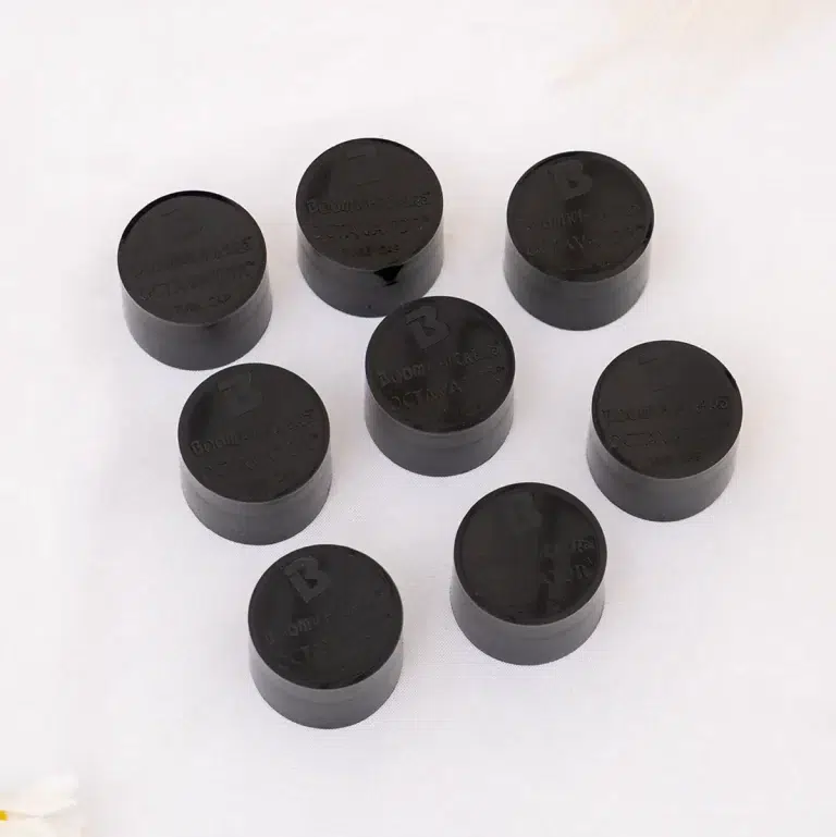 Nine black plastic bottle caps arranged in a loose circle on a white surface, with embossed lettering on the tops. ขายราคาพิเศษ