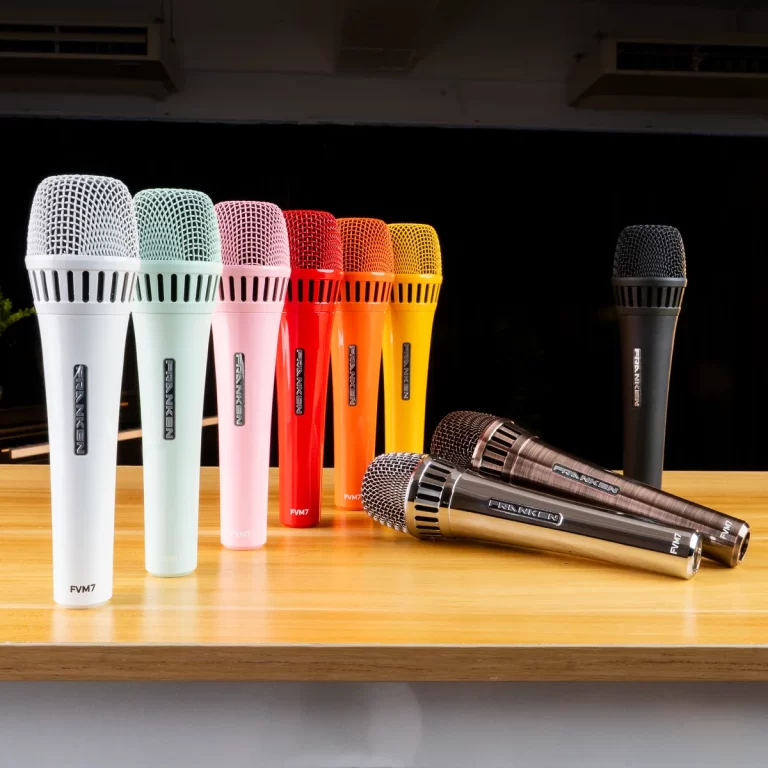 Row of colorful handheld microphones lined up on a wooden table, white to pink, red, orange, yellow, and a few metallic ones with brand labels visible ขายราคาพิเศษ