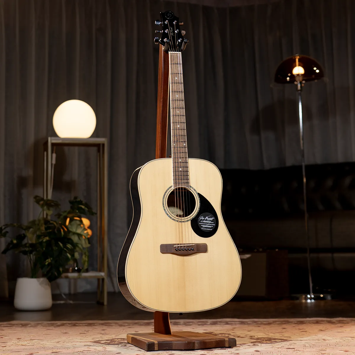 Acoustic guitar on a wooden stand in a softly lit studio, with a round lamp and plants in the background.