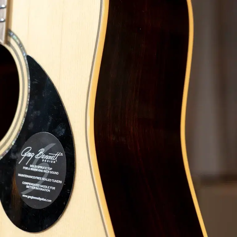Close-up of an acoustic guitar body: light spruce top, dark back, circular pickguard area near the soundhole with a brand badge visible. ขายราคาพิเศษ