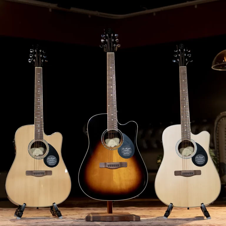 Three acoustic guitars on stands on a wooden stage, with a sunburst-finished guitar in the center flanked by two lighter guitars on either side. ขายราคาพิเศษ