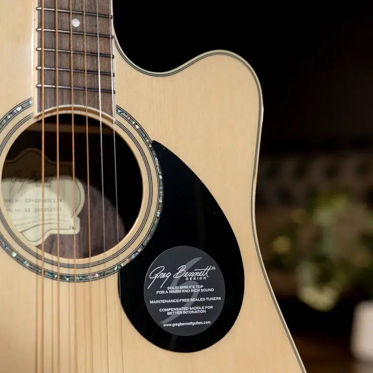 Close-up of an acoustic guitar's body and soundhole, with a black pickguard and a circular 'Greg Bennett Design' sticker near the soundhole badge ขายราคาพิเศษ
