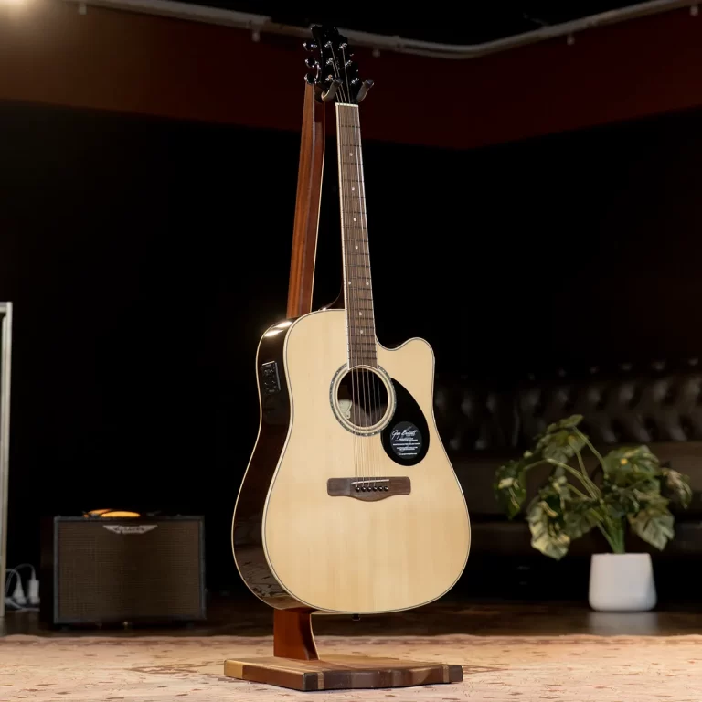 Light-colored acoustic guitar on a wooden stand in a dim stage setting, with a small amplifier and potted plant in the background. ขายราคาพิเศษ