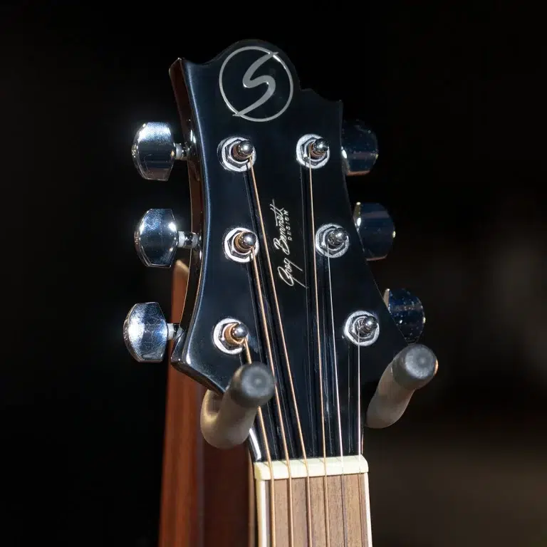 Close-up of a dark guitar headstock with six tuning pegs, strings, and a circular logo at the top, against a dark background. ขายราคาพิเศษ