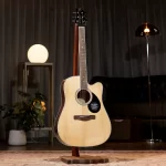 Light-colored acoustic guitar on a stand in a warmly lit room with a planter and lamps. ลดราคาพิเศษ