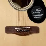 Close-up of an acoustic guitar's bridge and strings on a light wood body, with a black circular label near the soundhole. ขายราคาพิเศษ