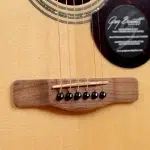 Close-up of an acoustic guitar bridge with six black bridge pins and a saddle wire, on a light wood body surface ขายราคาพิเศษ