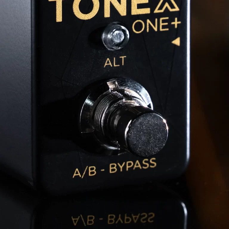 Close-up of a black guitar effects pedal front panel showing the ALT switch and the large A/B bypass footswitch with gold text reads 'TONE ONE+'. ขายราคาพิเศษ