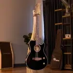 Black acoustic guitar on a stand in a cozy studio, with a wooden shelf and amplifier in the background ขายราคาพิเศษ