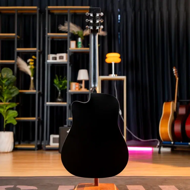 Black acoustic guitar on a stand in a studio showroom with shelves, plants, and other guitars in the background. ขายราคาพิเศษ