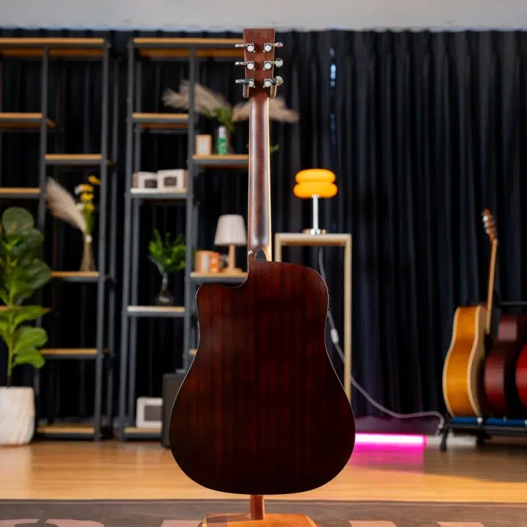 Dark brown acoustic guitar on a stand, facing away, in a shop/studio with shelves, plants, and other guitars in the background ขายราคาพิเศษ