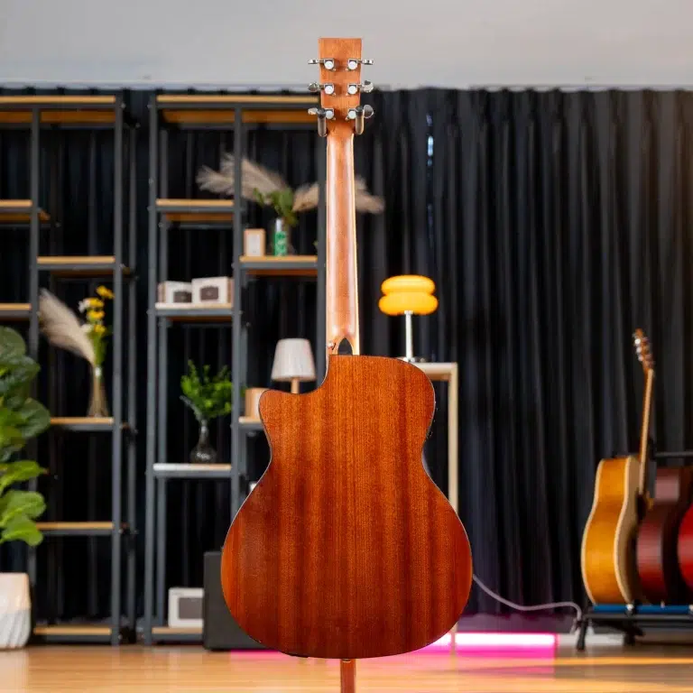 Front-facing view of a glossy brown acoustic guitar standing upright on a stand in a showroom, with other guitars and shelving in the background. ขายราคาพิเศษ