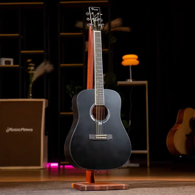 Black acoustic guitar on a wooden stand centered on a dimly lit stage, with amplifier and other instruments in the background. ขายราคาพิเศษ