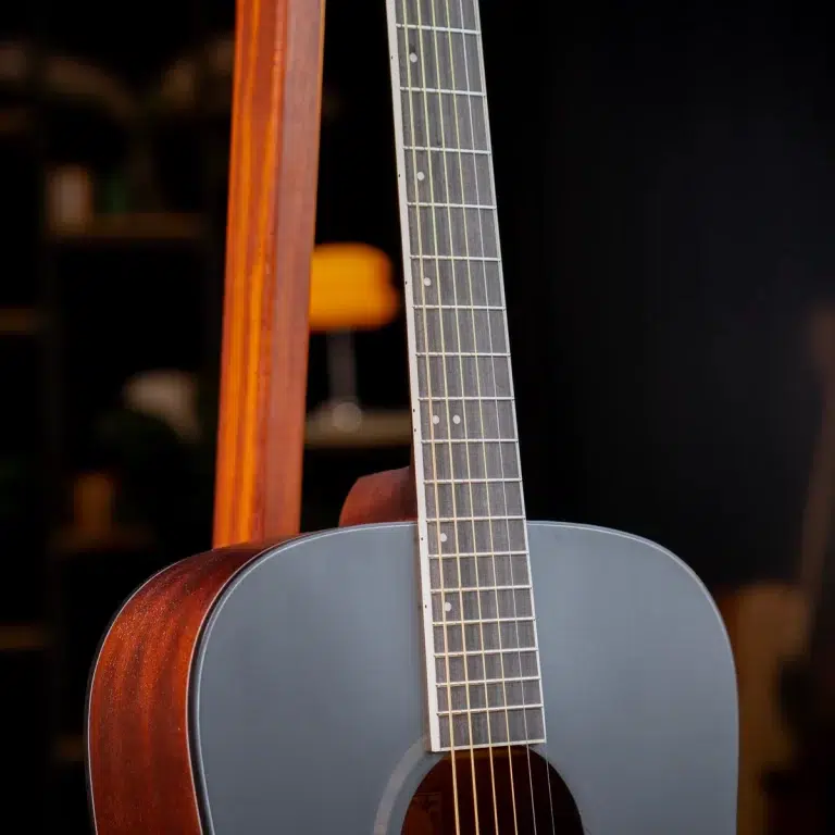 Close-up of a blue-gray acoustic guitar showing the neck, frets, and strings against a dark background. A wooden frame supports the guitar in the foreground. ขายราคาพิเศษ