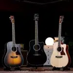 Three acoustic guitars on stands in a dark room, with a sunburst, black, and natural finish from left to right, and a warm round lamp behind them. ลดราคาพิเศษ