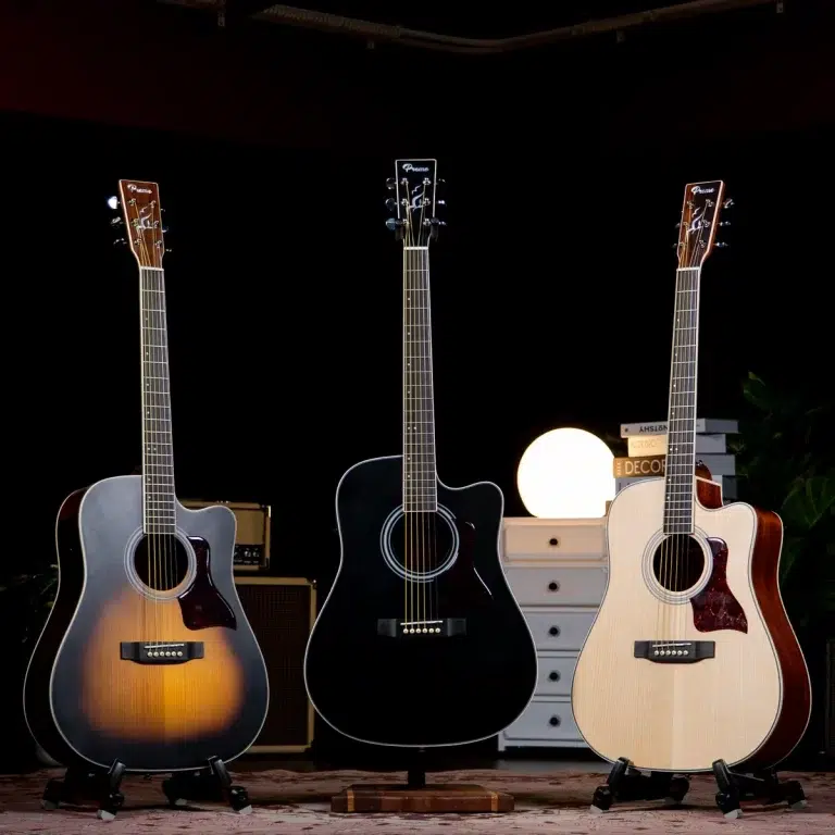 Three acoustic guitars on stands in a dark room, with a sunburst, black, and natural finish from left to right, and a warm round lamp behind them. ขายราคาพิเศษ