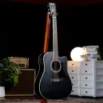 Black acoustic guitar on a wooden stand in a home studio setup with an amp, plants, and white drawers in the background ขายราคาพิเศษ