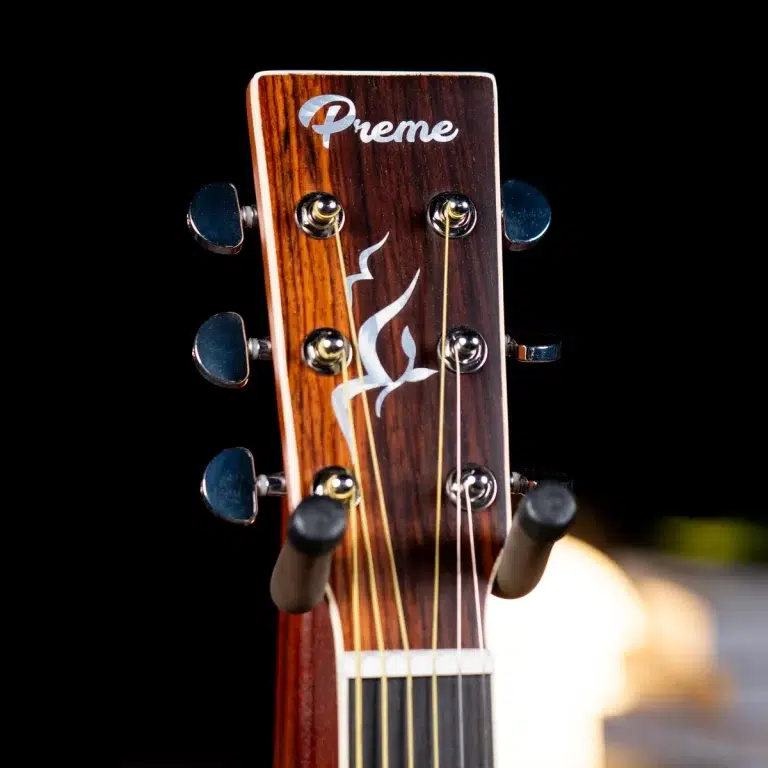 Close-up of a wooden guitar headstock with six tuning pegs and strings, featuring a white 'Preme' logo and flame design against a black background. ขายราคาพิเศษ