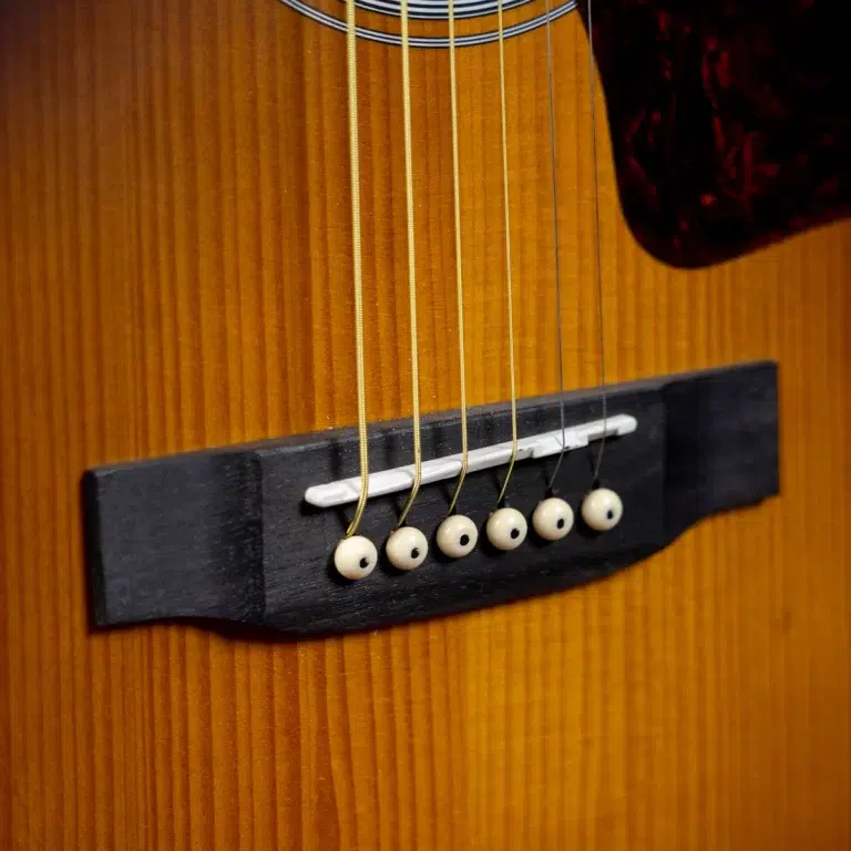 Close-up of an acoustic guitar bridge with six white bridge pins and a saddle on a wood body, showing six strings anchored downwards. ขายราคาพิเศษ