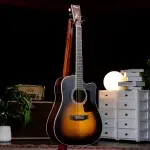 Sunburst acoustic guitar on a stand in a dim studio setting with warm lighting and gear in the background ขายราคาพิเศษ