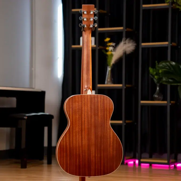 Back view of a brown acoustic guitar standing upright in a room with a black piano on the left and decorative shelves in the background. ขายราคาพิเศษ