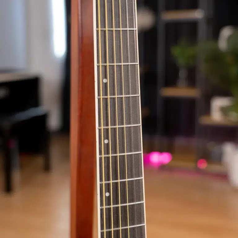 Close-up of a guitar neck with frets and strings in focus, blurred living room background behind it. ขายราคาพิเศษ