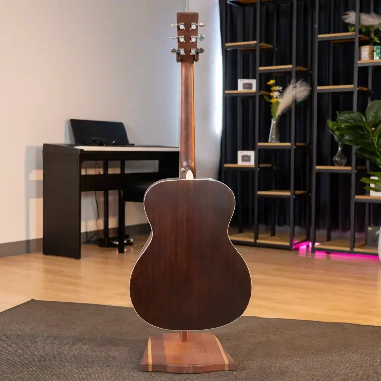 Back of a dark wood acoustic guitar on a stand in a showroom or studio, with a keyboard and shelves in the background. ขายราคาพิเศษ